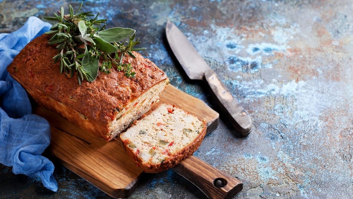 Draufsicht: Auf einem Untergrund aus Metall liegt ein Holzbrettchen mit einem angeschnittenen Schinken-Oliven-Kuchen. Auf dem Küchen liegen frische Kräuter. Und neben dem Brettchen ein großes, altes Küchenmesser.