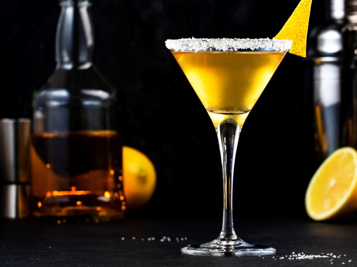 Sidecar Cocktail in Cocktailschale mit Zitronenzeste vor schwarzem Hintergrund. Im Hintergrund außerdem eine Flasche Cognac, Barmaß, Zitronen und Shaker. Frontalansicht.