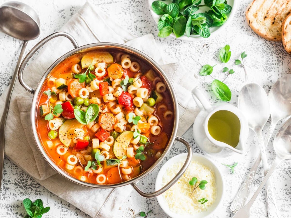 Sommerliche Minestrone: ein Topf mit Suppe, darin Nudeln und Gemüse, drumherum allerlei Zutaten.