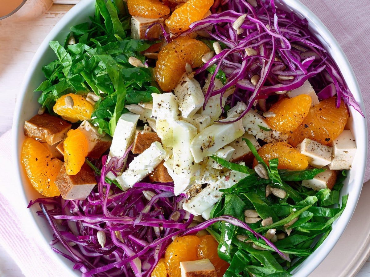 Spinat-Salat mit Ziegenkäse und Mandarinen in einer weißen Schüssel vor hellem Hintergrund in der Draufsicht.