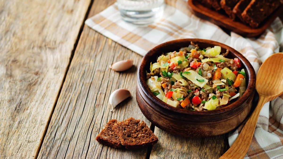 Spitzkohleintopf mit Hackfleisch und Kartoffeln in Tonschale auf Holz. Im Bild Brot und Knoblauch. Draufsicht.