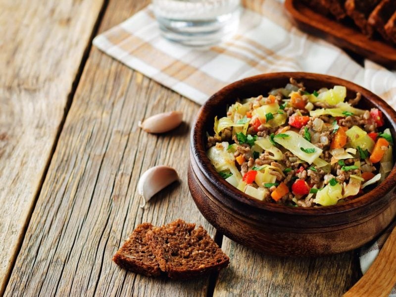 Spitzkohleintopf mit Hackfleisch und Kartoffeln in Tonschale auf Holz. Im Bild Brot und Knoblauch. Draufsicht.