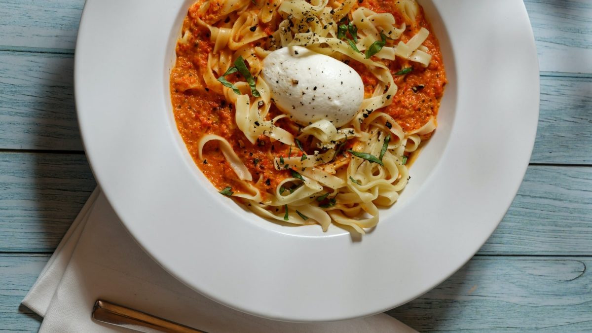 Draufsicht: Türkisfarbener Holzuntergrund mit einem tiefen Teller im Bildzentrum. Darauf Tagliatelle mit Paprikasauce und Burrata.