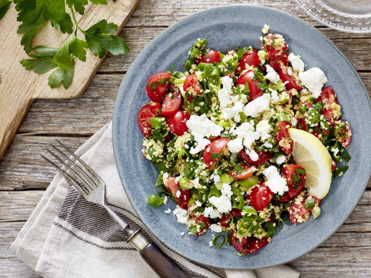 Draufsicht: Holzuntergrund mit einem Holzbrett oben links, einer beigen Stoffservierte unten und einem blauen Teller im Bildzentrum. Darauf ein Tomaten-Amaranth-Salat mit Petersilie.