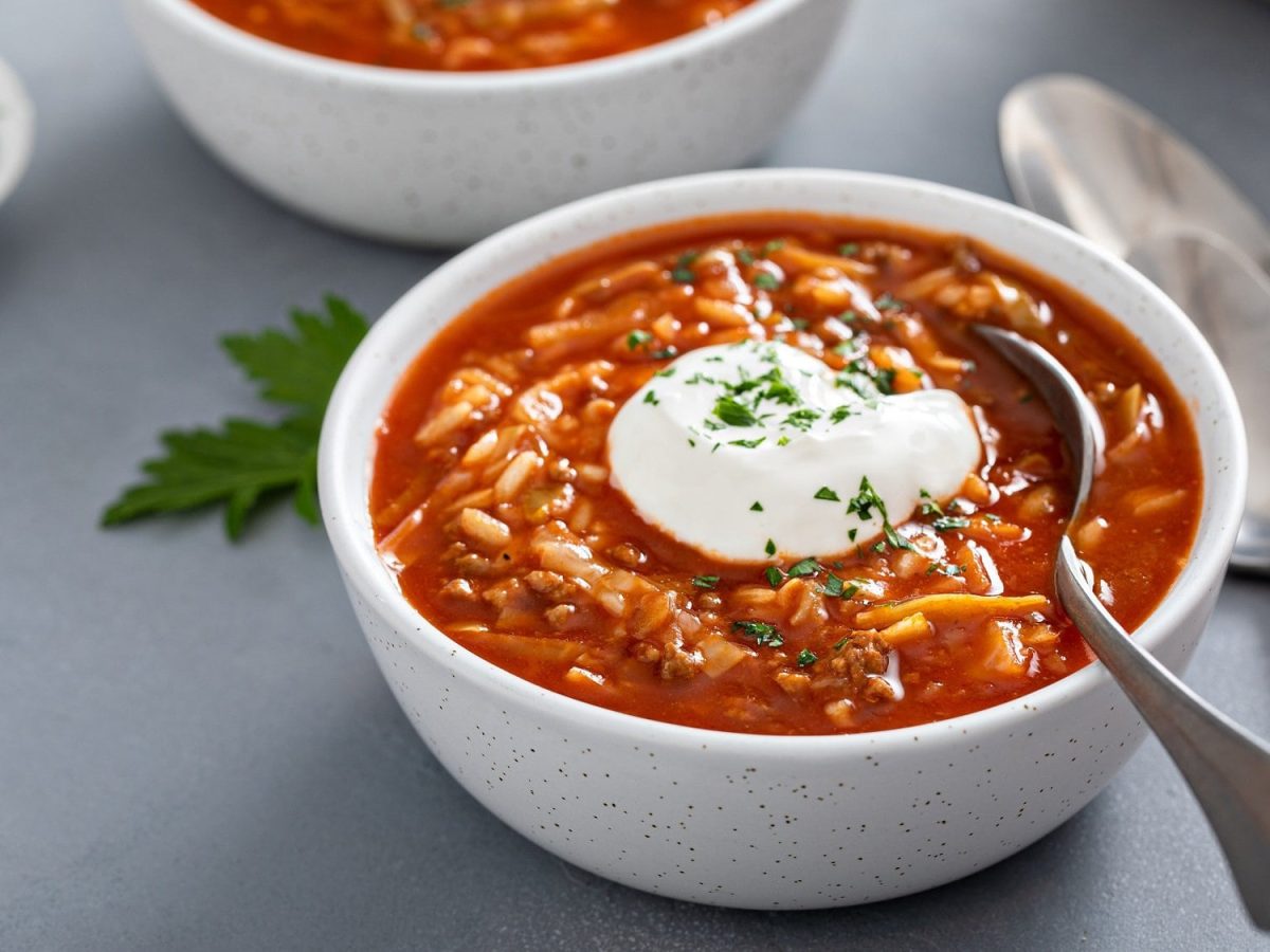 Eine Schüssel Tomaten-Reis-Suppe mit einem Klecks Crème fraîche und einem Löffel darin, daneben liegen weitere Löffel.