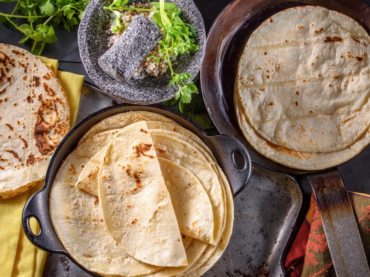 Tortillas aus Maismehl und aus Weizenmehl auf Tellern und in Pfannen verteilt. Im Bild Füllungen und Toppings. Links und rechts Tücher im mexikanischen Stil. Draufsicht.