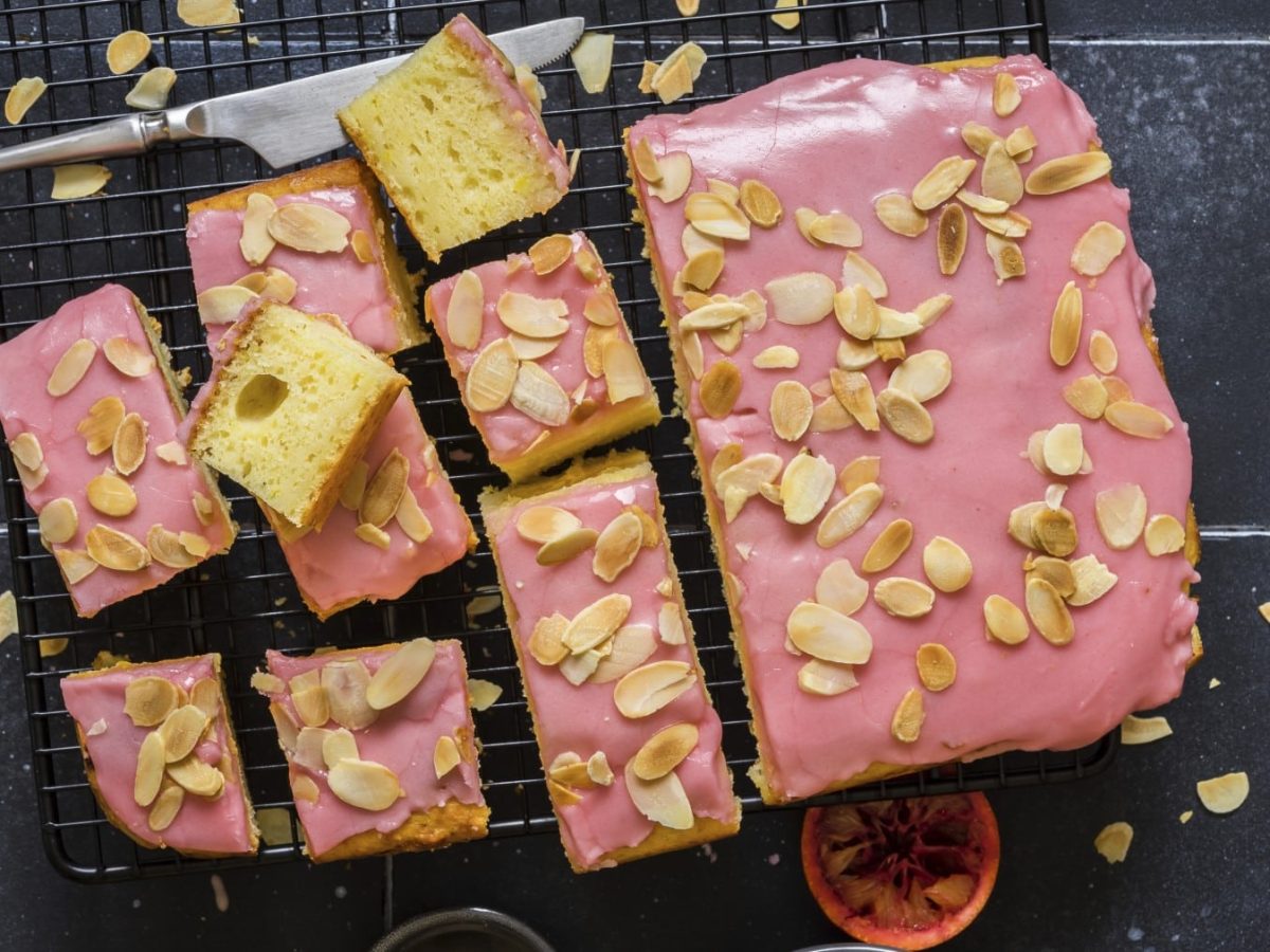 Zitronen-Ricotta-Kuchen mit Blutorangenglasur und Mandelblättchen angeschnitten auf einem Gitter in der Draufsicht.