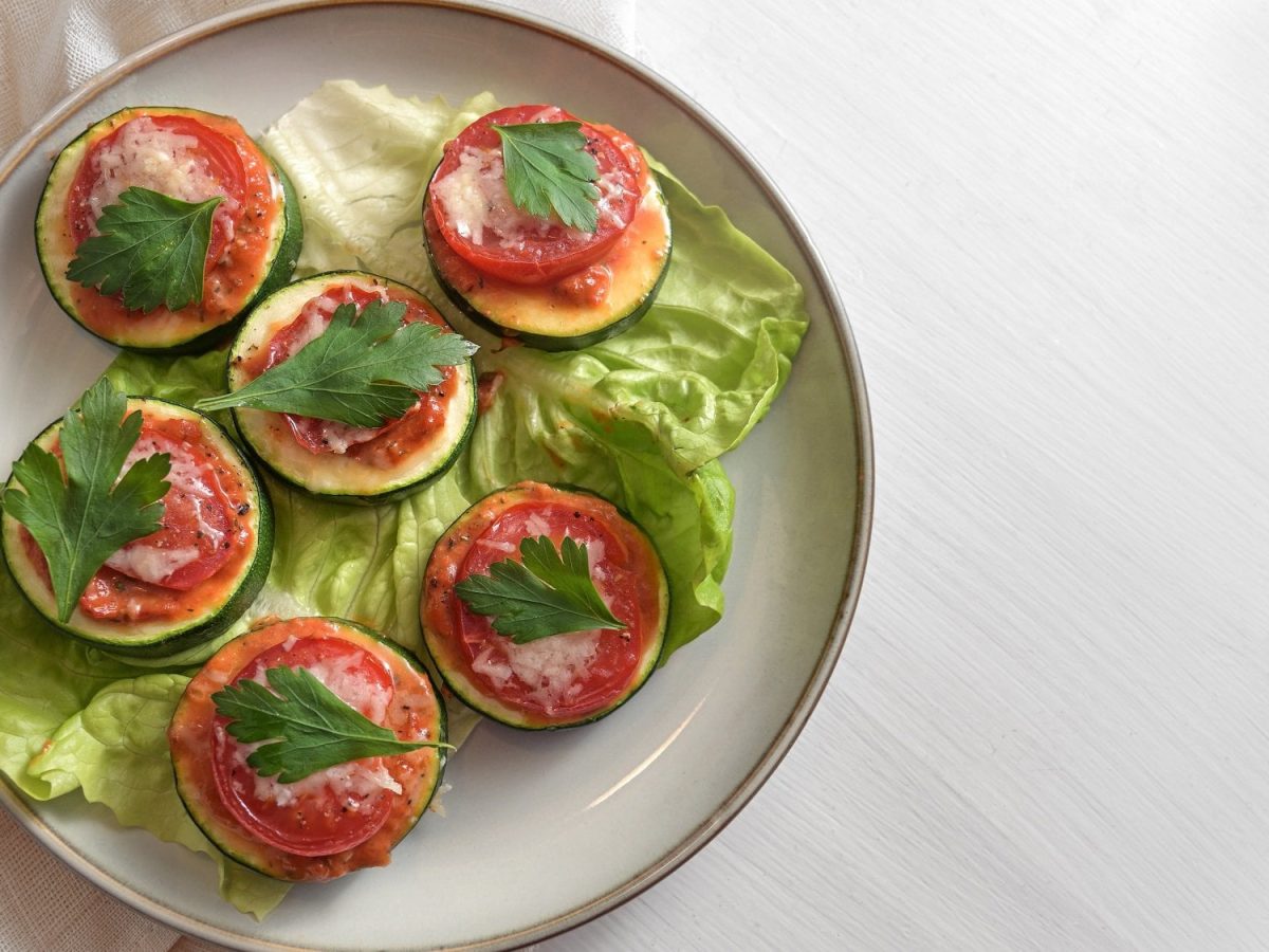 Mehrere Zucchini-Bruschetta-Häppchen mit einer Tomate und Petersilie obenauf auf einem Teller, Draufsicht.