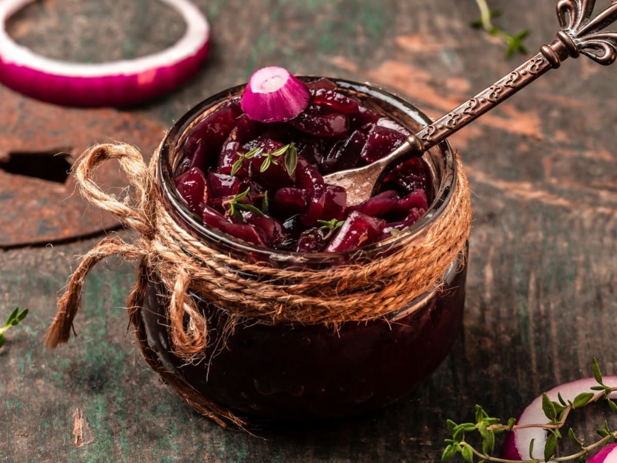 Schräge Ansicht: Holztisch auf dem ein kleines Schraubglas steht. Darin enthalten würziges Zwiebel-Chutney aus roten Zwiebeln, mit Thymian.