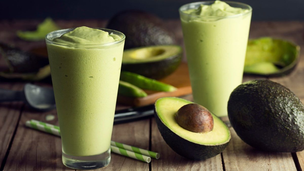 Zwei Gläser mit dem grünen Avocado-Mango-Lassi auf einem Holztisch mit halbierter Avocado im Hintergrund.