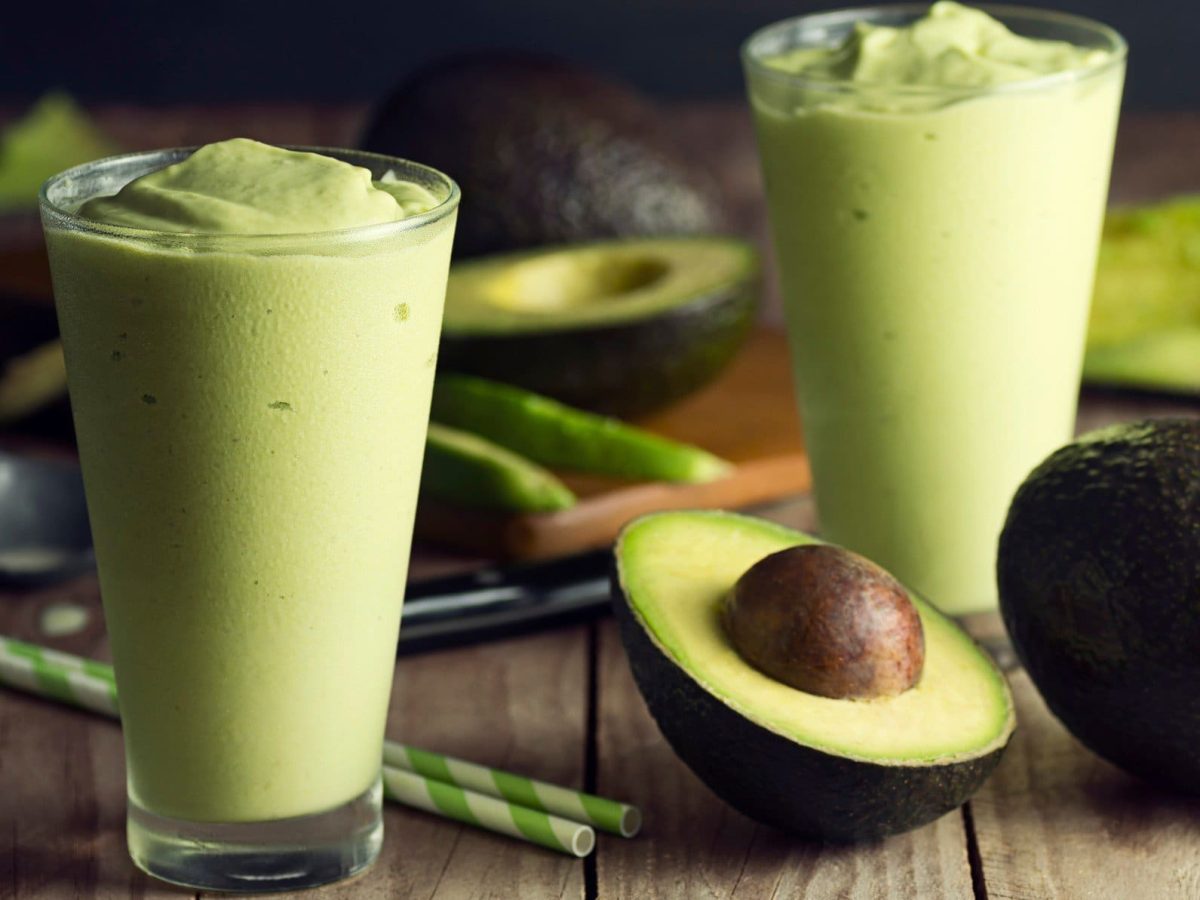 Zwei Gläser mit dem grünen Avocado-Mango-Lassi auf einem Holztisch mit halbierter Avocado im Hintergrund.