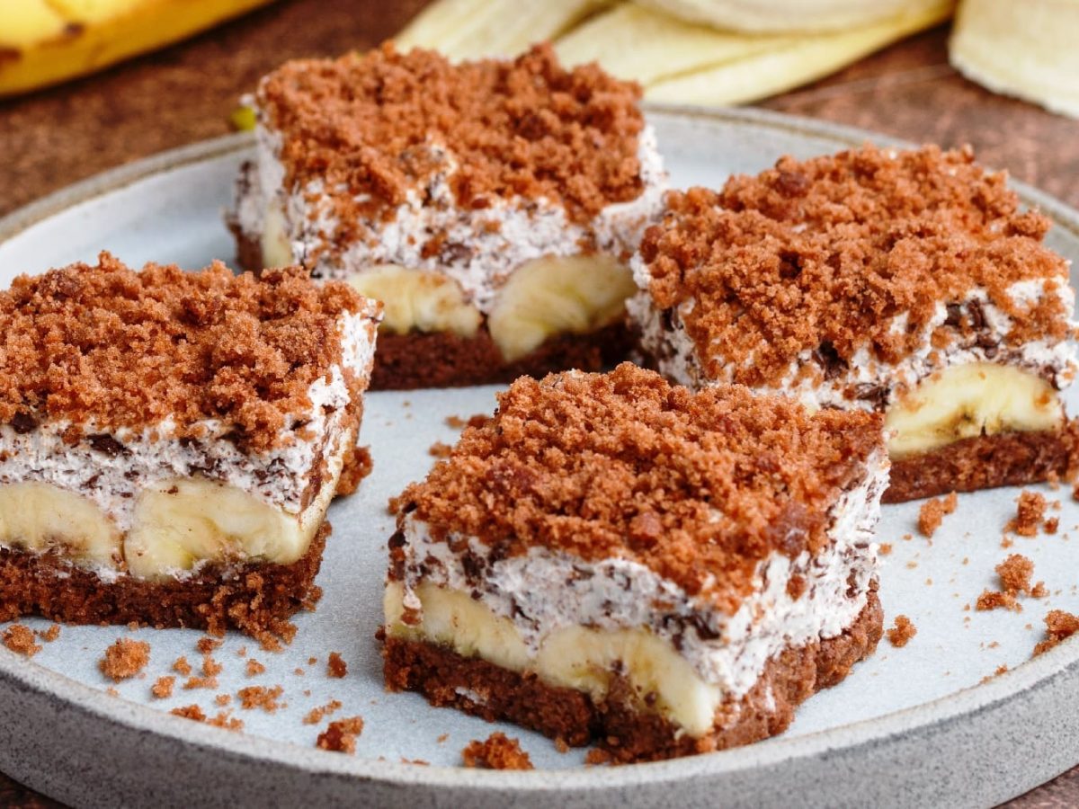 Vier Stücken von dem Bananenkuchen auf einem dunkelbraunem Tisch. Im Hintergrund eine geschälte Banane und ein paar Schokostücken.