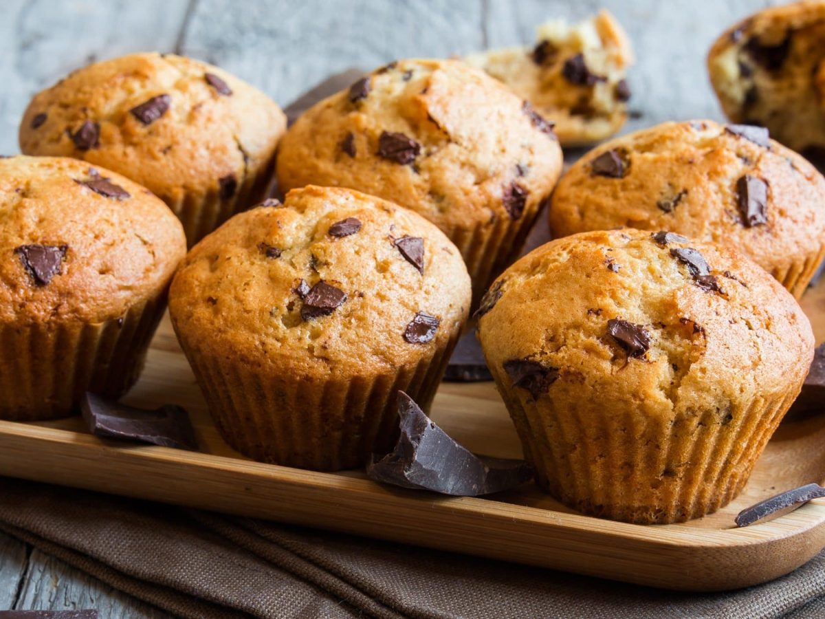 6 Bananenbrot-Muffins mit Schokodrops auf einem Holztablett, im Hintergrund noch ein Muffins.