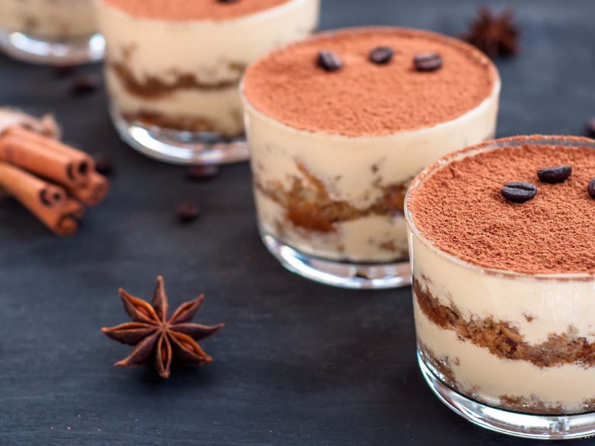 Vier Gläser Biscoff-Tiramisu mit Kekskrümeln, daneben Sternanis und Zimtstangen.