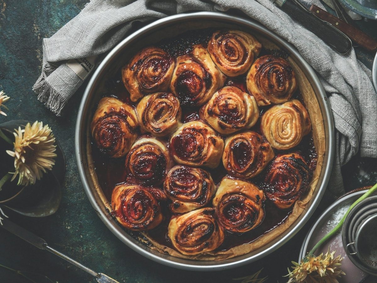 Draufsicht: Dunkler Untergrund auf dem mittig eine Backform mit buttrigen Blaubeermarmeladenschnecken steht. Darum verteilt ein paar Backutensilien und Blumen.