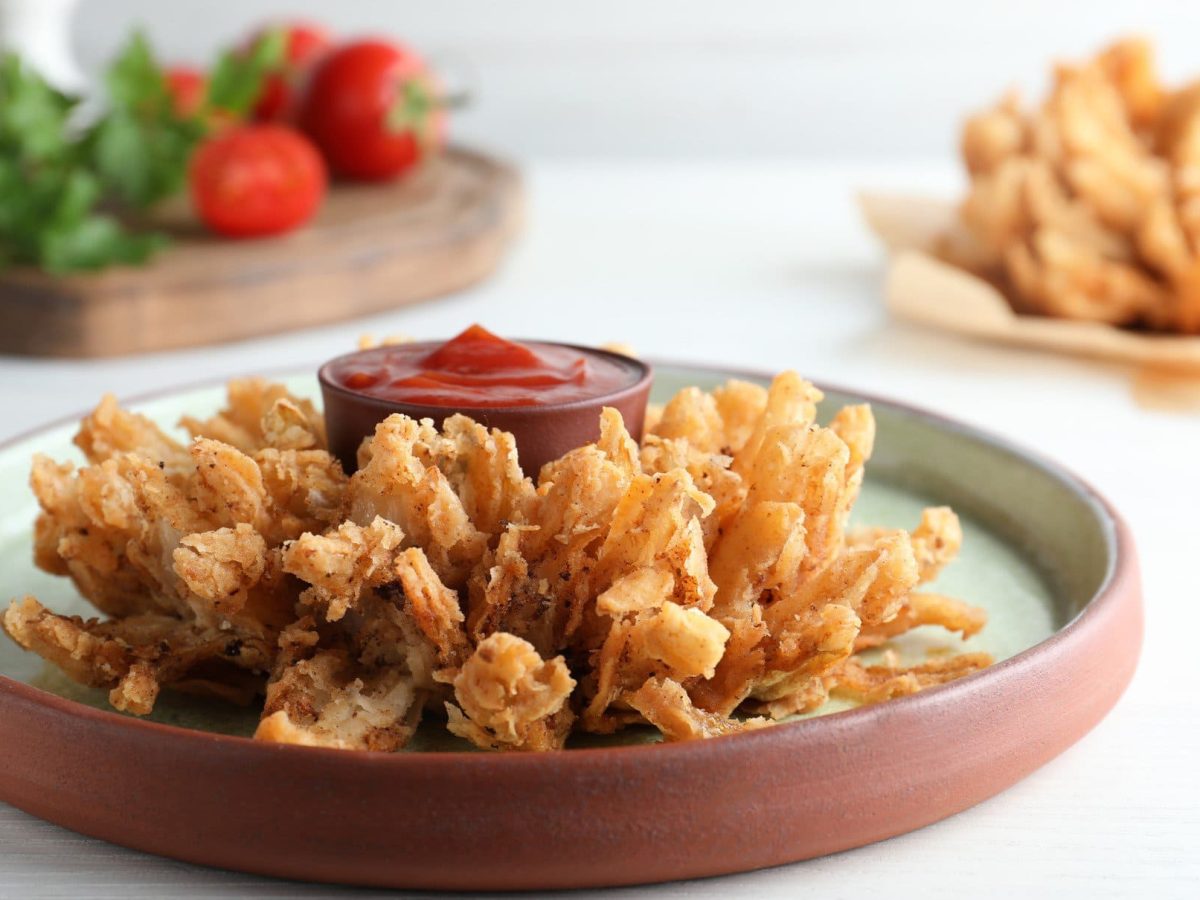 Zwei Blooming Onion frittierte Zwiebelblume auf Tellern und hellem Untergrund. In der Mitte eine Schale mit rotem Dip. Im Hintergrund Salat auf Holzbrett. Frontalansicht.