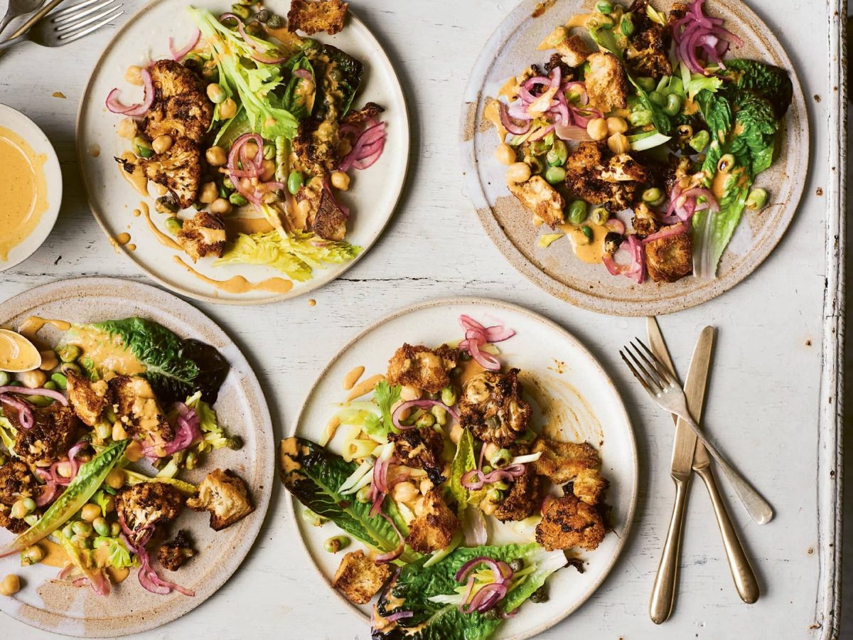 Vier Teller mit de Blumenkohl-Caesar-Salad auf einem hellen Holztisch.