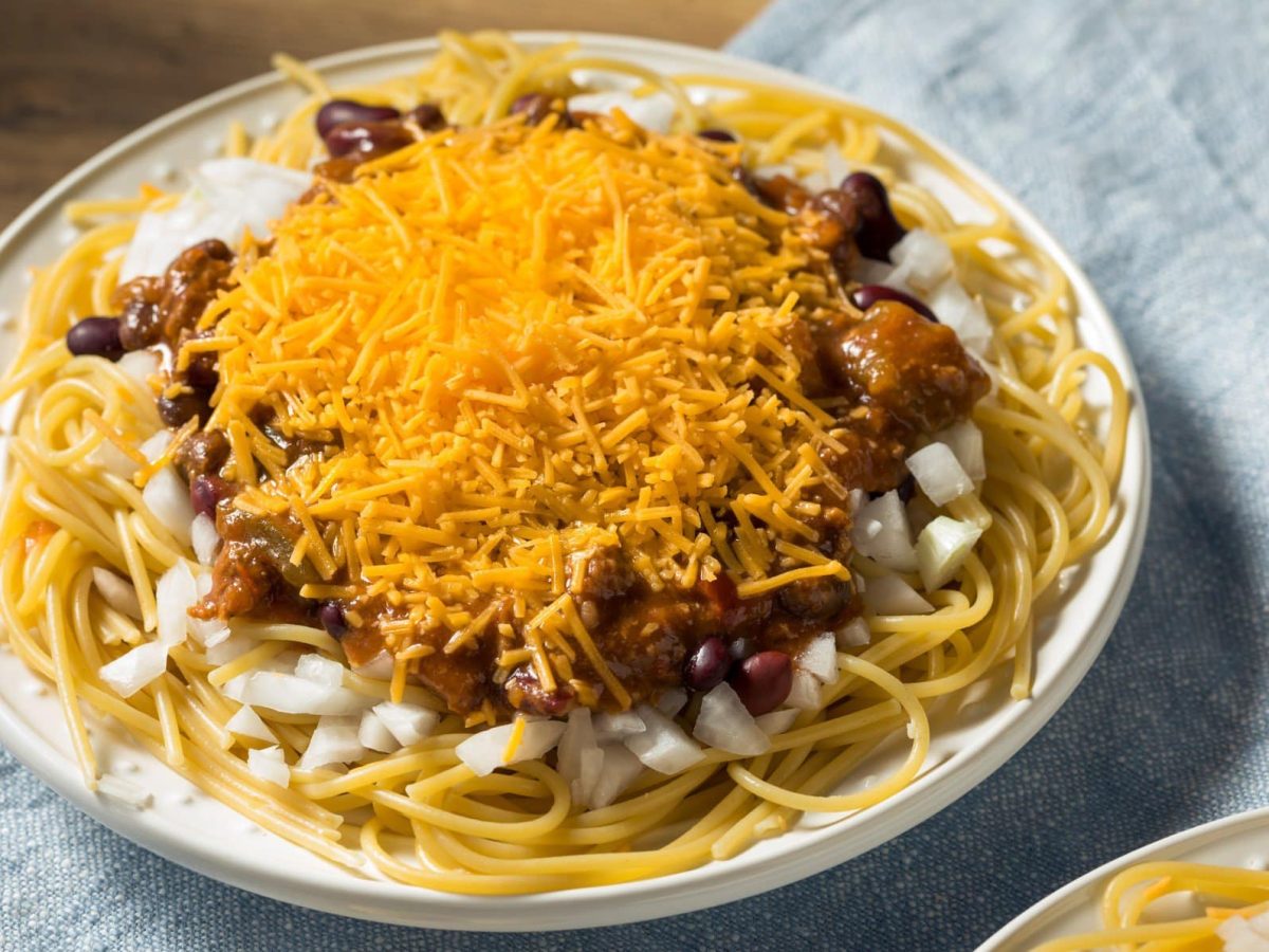 Cincinnati Chili mit Cheddar auf Pastateller, blauem Tuch und Holz. Unten eine weitere Portion angedeutet. Draufsicht.