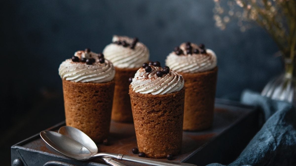 Dunkler Hintergrund. Vier Cookie-Cups nach Tiramisu-Art auf einem dunklen Koffer. Davor zwei Silberlöffel. Im rechten Bildrand eine blaue Stoffservierte und im Hintergrund Schleiherkraut.