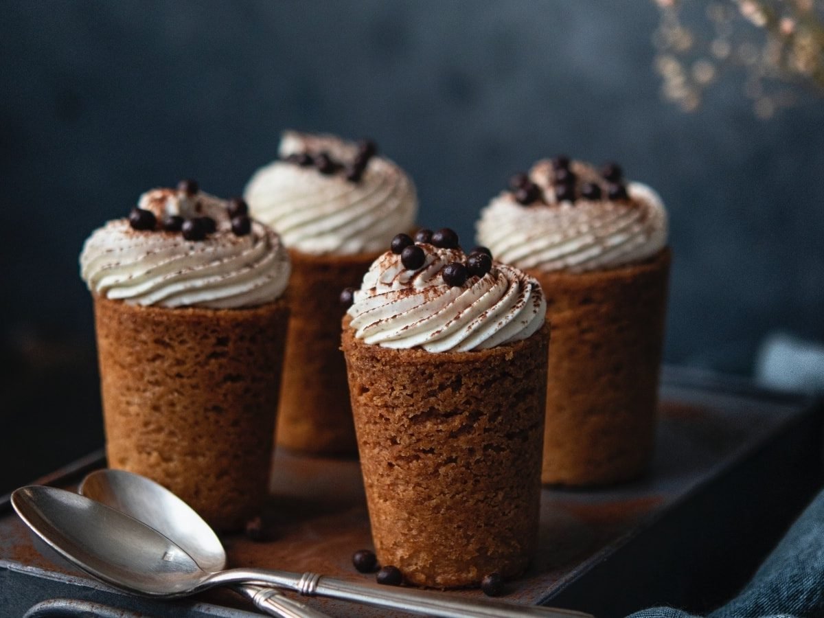 Dunkler Hintergrund. Vier Cookie-Cups nach Tiramisu-Art auf einem dunklen Koffer. Davor zwei Silberlöffel. Im rechten Bildrand eine blaue Stoffservierte und im Hintergrund Schleiherkraut.