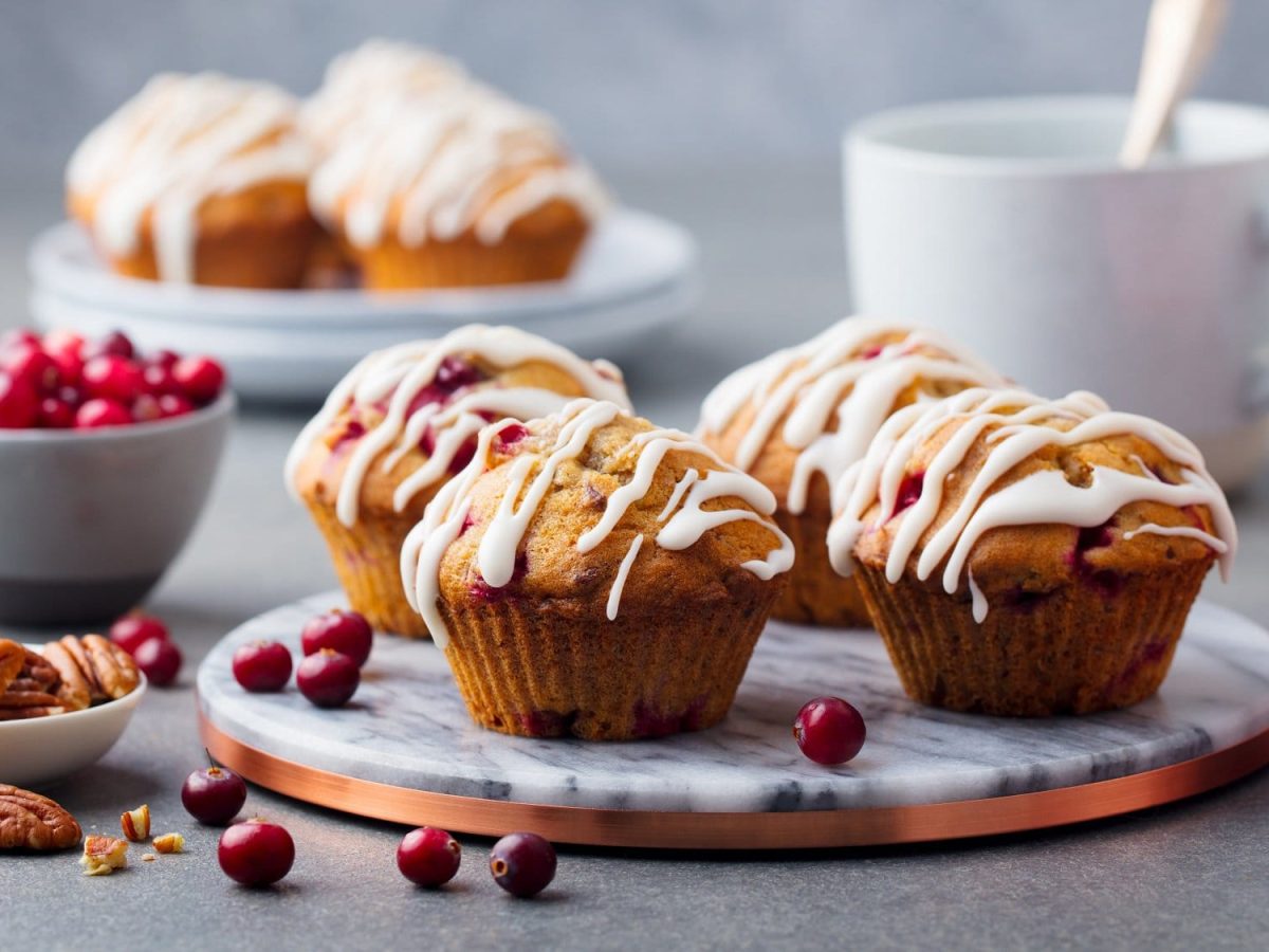 Cranberry-Muffins auf einem Marmor-Tablett, daneben eine Schale mit Cranberrys und Nüssen.