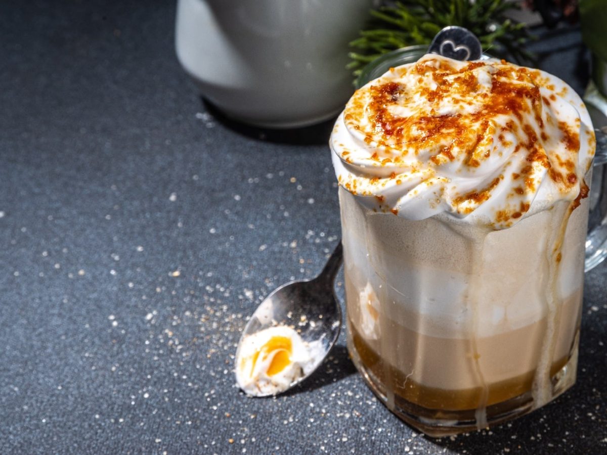 Eine Tasse mit dem Crème-Brûlée-Latte auf einem dunklen Untergrund. Im Hintergrund unscharf Kaffeeequipment.