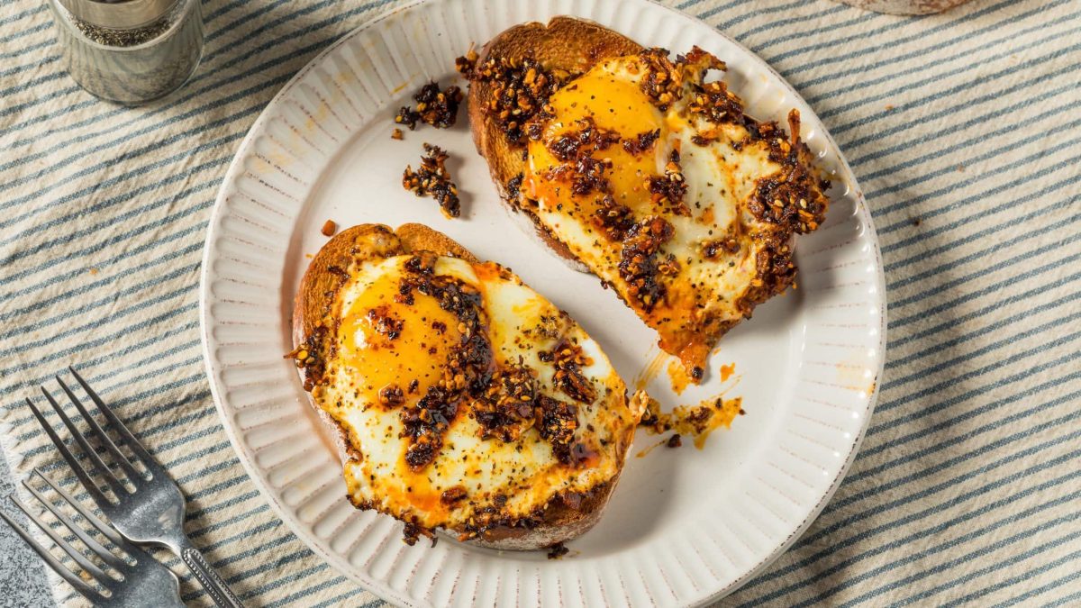 Zwei Crispy Chili Oil Egg Toast auf weißem Teller und hellem Untergrund. Daneben Besteck und Salzstreuer. Draufsicht.