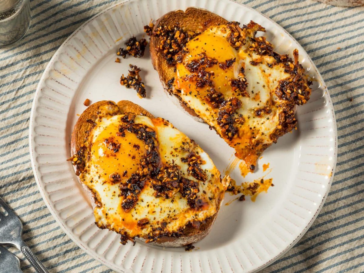 Zwei Crispy Chili Oil Egg Toast auf weißem Teller und hellem Untergrund. Daneben Besteck und Salzstreuer. Draufsicht.