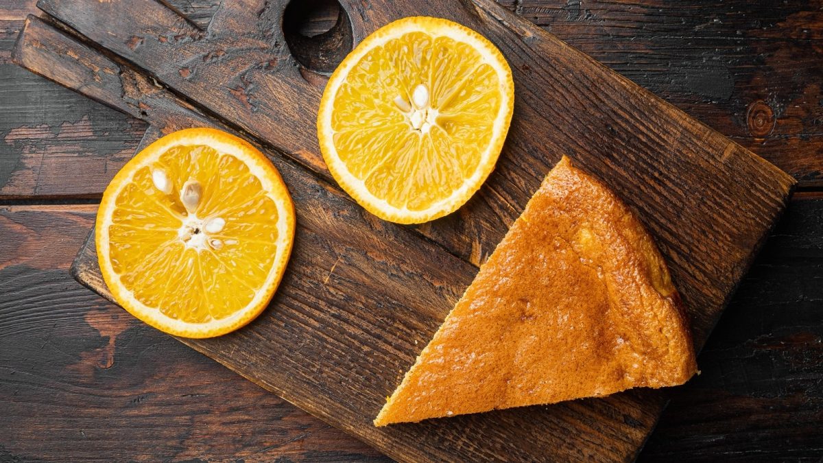 Eine Scheibe Orangen-Polentakuchen auf einem Holztisch neben Orangenscheiben.