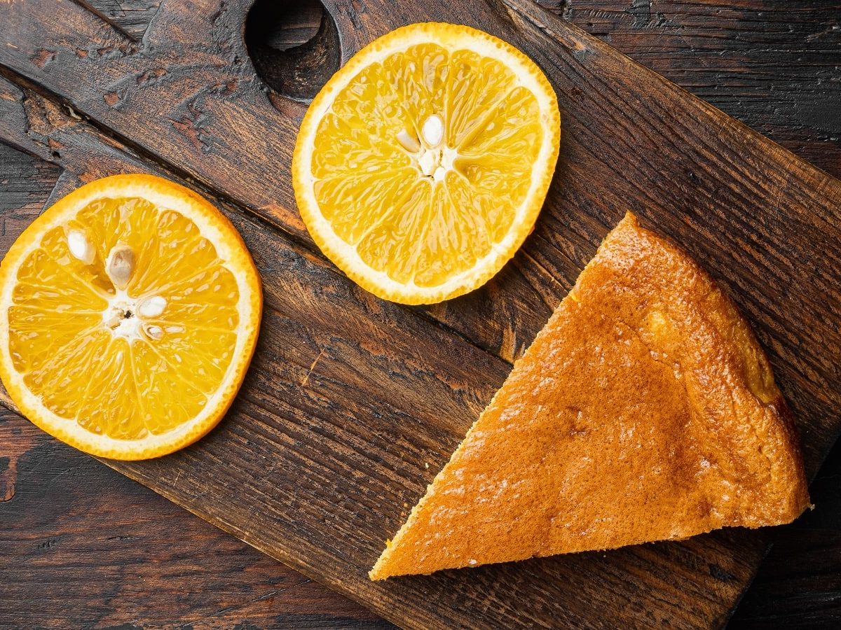 Eine Scheibe Orangen-Polentakuchen auf einem Holztisch neben Orangenscheiben.