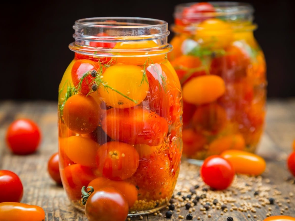 Nahaufnahme: Schwarzer Hintergrund, mit Holztisch auf dem zwei Schraubgläser mit eingelegten Tomaten in Honig-Sud stehen. Drumherum ein paar frische Tomaten.