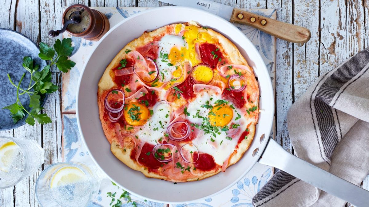 Draufsicht: Grauer Untergrund mit einer Stahlpfanne in der Bildmitte. Drumherum ein paar frische Kräuter, ein Messer, ein Küchenhandtuch. In der Pfanne eine Frühstückspizza mit Eiern und Kochschinken.