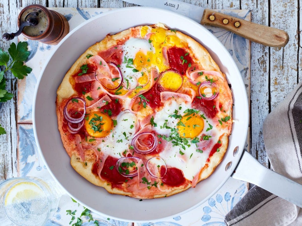 Draufsicht: Grauer Untergrund mit einer Stahlpfanne in der Bildmitte. Drumherum ein paar frische Kräuter, ein Messer, ein Küchenhandtuch. In der Pfanne eine Frühstückspizza mit Eiern und Kochschinken.