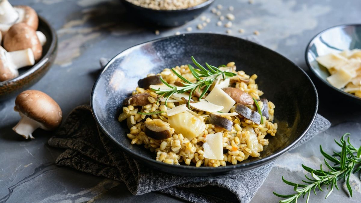 Dunkelgrauer Untergrund. In der Bildmitte ein Teller auf dem Gersotto mit Fenchel und gerösteten Pilzen ist. Darunter eine graue Stoffservierte. Um den Teller weitere Schüsseln mit frischen Pilzen, Parmesanhobeln und Rosmarin.