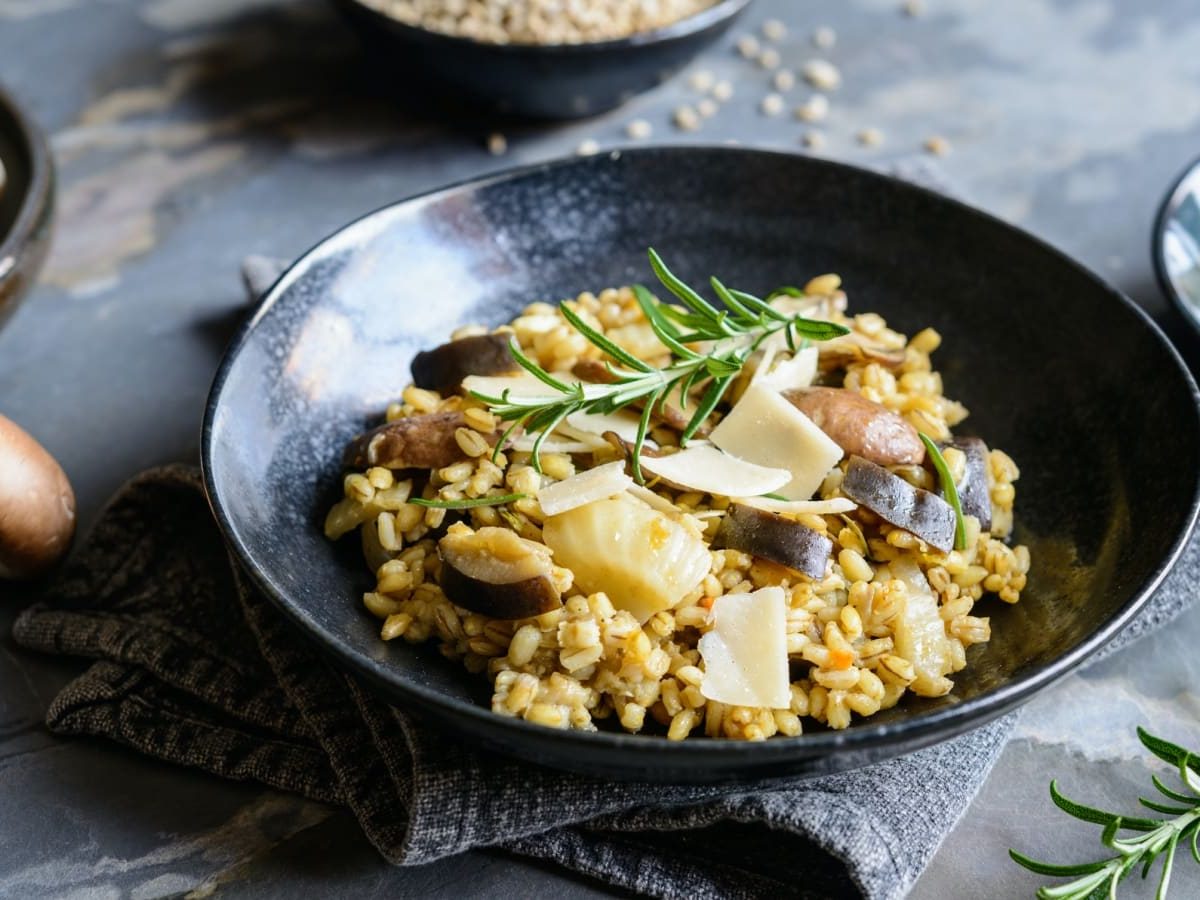 Dunkelgrauer Untergrund. In der Bildmitte ein Teller auf dem Gersotto mit Fenchel und gerösteten Pilzen ist. Darunter eine graue Stoffservierte. Um den Teller weitere Schüsseln mit frischen Pilzen, Parmesanhobeln und Rosmarin.