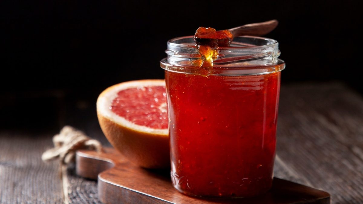 Schwarzer Hintergrund mit dunklem Holztisch. Darauf ein kleines Holzbrett auf dem ein Glas mit Grapefruit-Vanille-Gelee und einer aufgeschnittenen Grapefruit im Hintergrund.