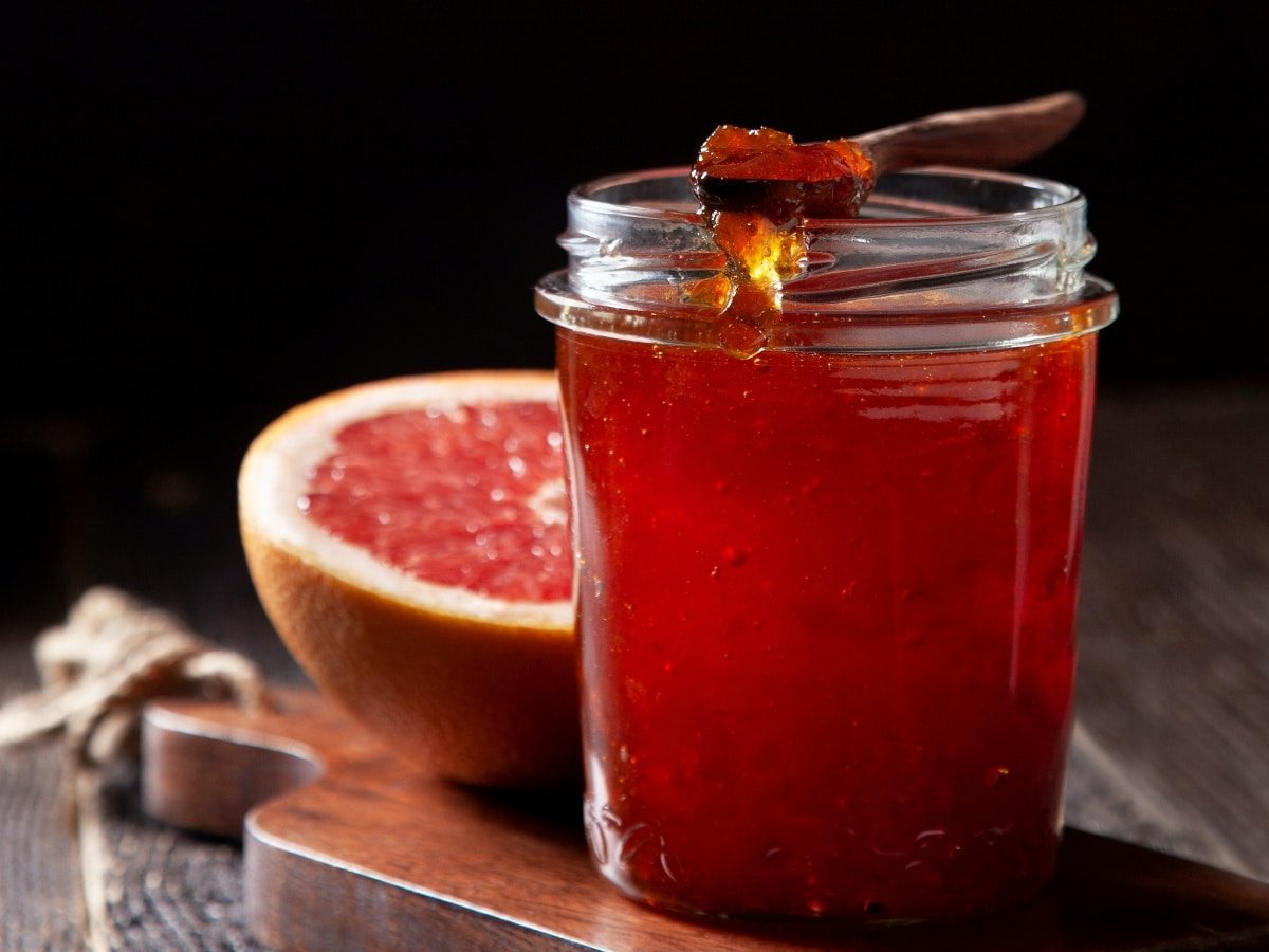 Schwarzer Hintergrund mit dunklem Holztisch. Darauf ein kleines Holzbrett auf dem ein Glas mit Grapefruit-Vanille-Gelee und einer aufgeschnittenen Grapefruit im Hintergrund.