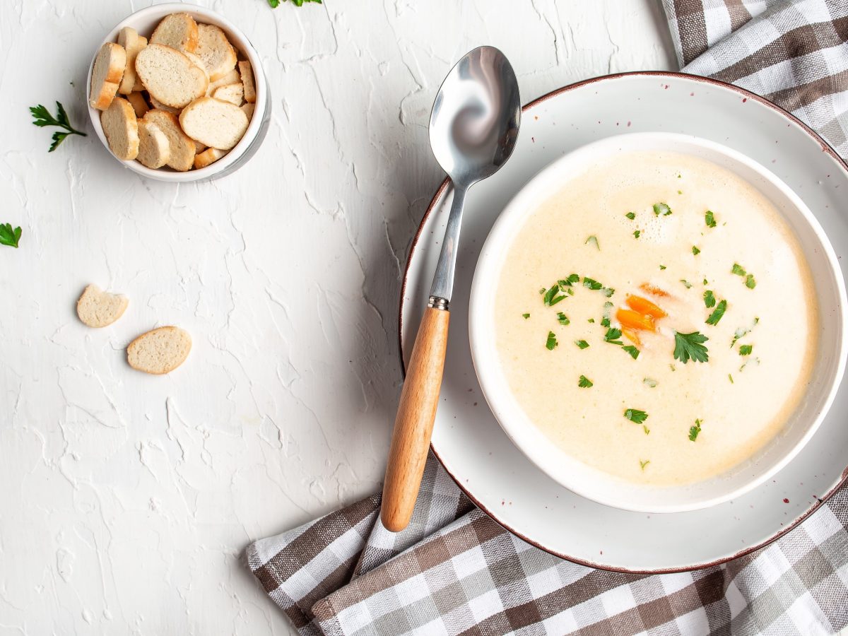 Draufsicht: eine Feine Hähnchen-Cremesuppe sowie ein Teller mit Croûtons und ein kariertes Küchentuch stehen auf einer hellen Tischplatte.