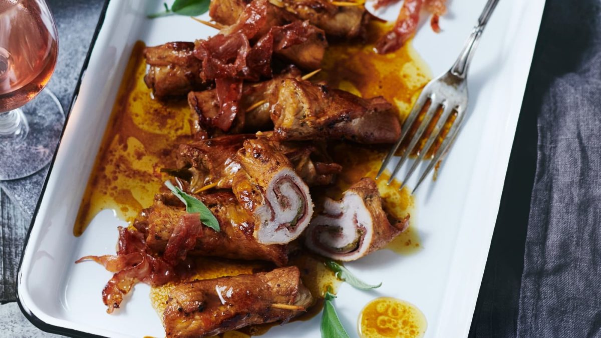 Draufsicht: Dunkler Untergrund mit einem weißen Teller auf dem Involtini alla Siciliana serviert werden. Links daneben steht ein Glas Roséweine.