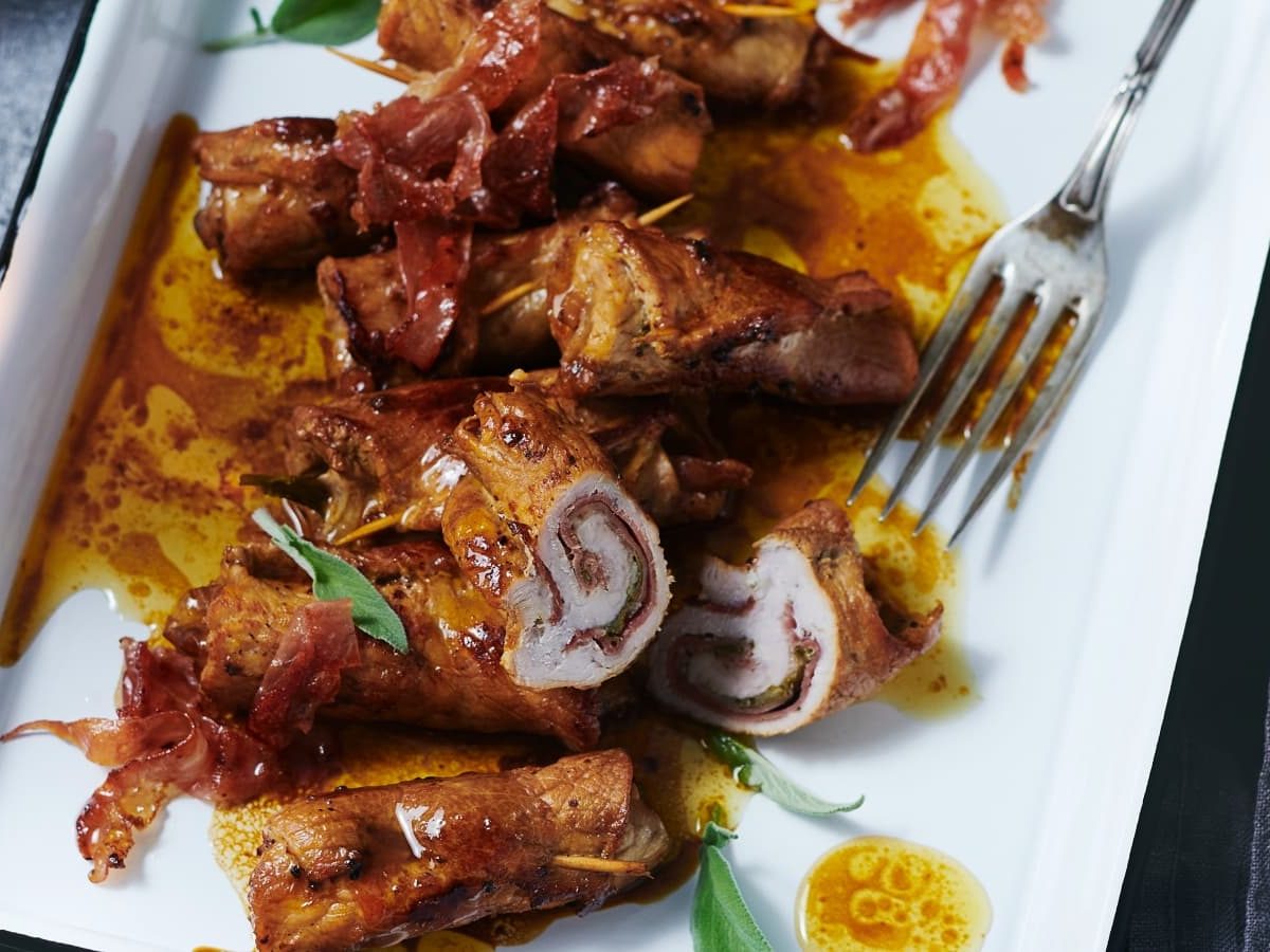 Draufsicht: Dunkler Untergrund mit einem weißen Teller auf dem Involtini alla Siciliana serviert werden. Links daneben steht ein Glas Roséweine.