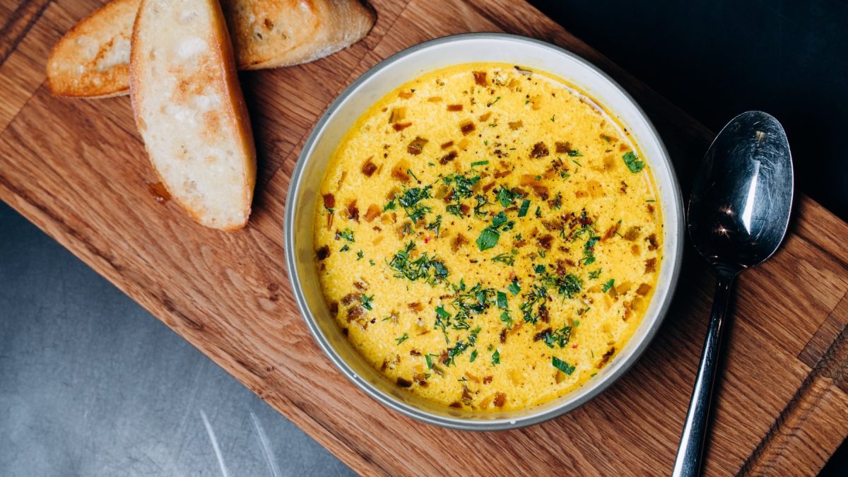 Ein Teller mit der Käsecremesuppe auf einem Holzbrett mit zwei Brotscheiben und dunklem Untergrund.