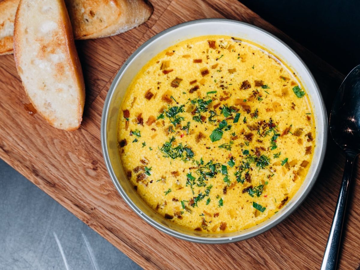 Ein Teller mit der Käsecremesuppe auf einem Holzbrett mit zwei Brotscheiben und dunklem Untergrund.