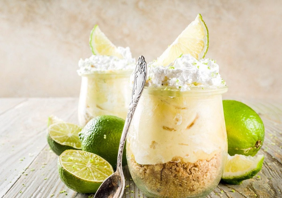 Frontalsicht: Zwei Kuchen im Glas, beziehungsweise Käse-Limetten-Kuchen, stehen auf einem Holztisch.. Drum herum liegen Limetten.