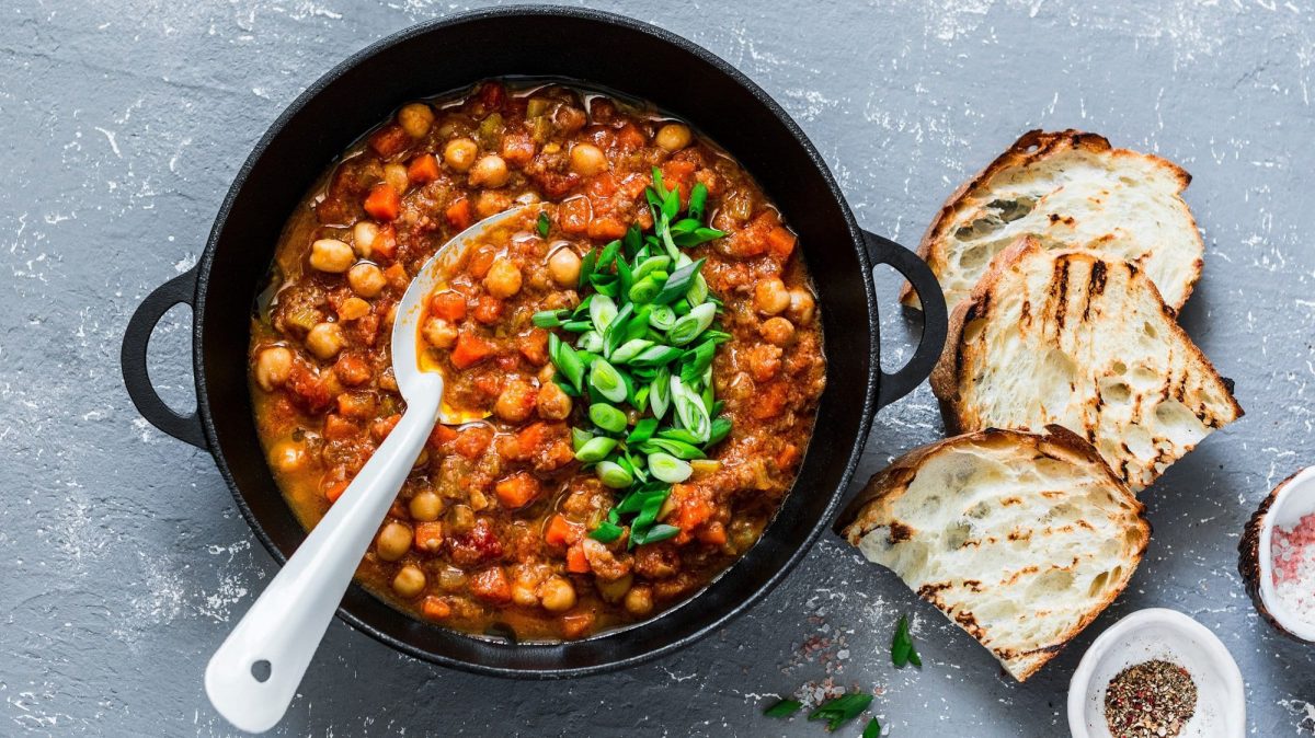 Ein Topf Kichererbsen-Chili mit Möhren, Tomaten und anderem Gemüse, außerdem Frühlingszwiebeln, daneben geröstete Brotscheiben und Schalen mit Gewürzen. Draufsicht.