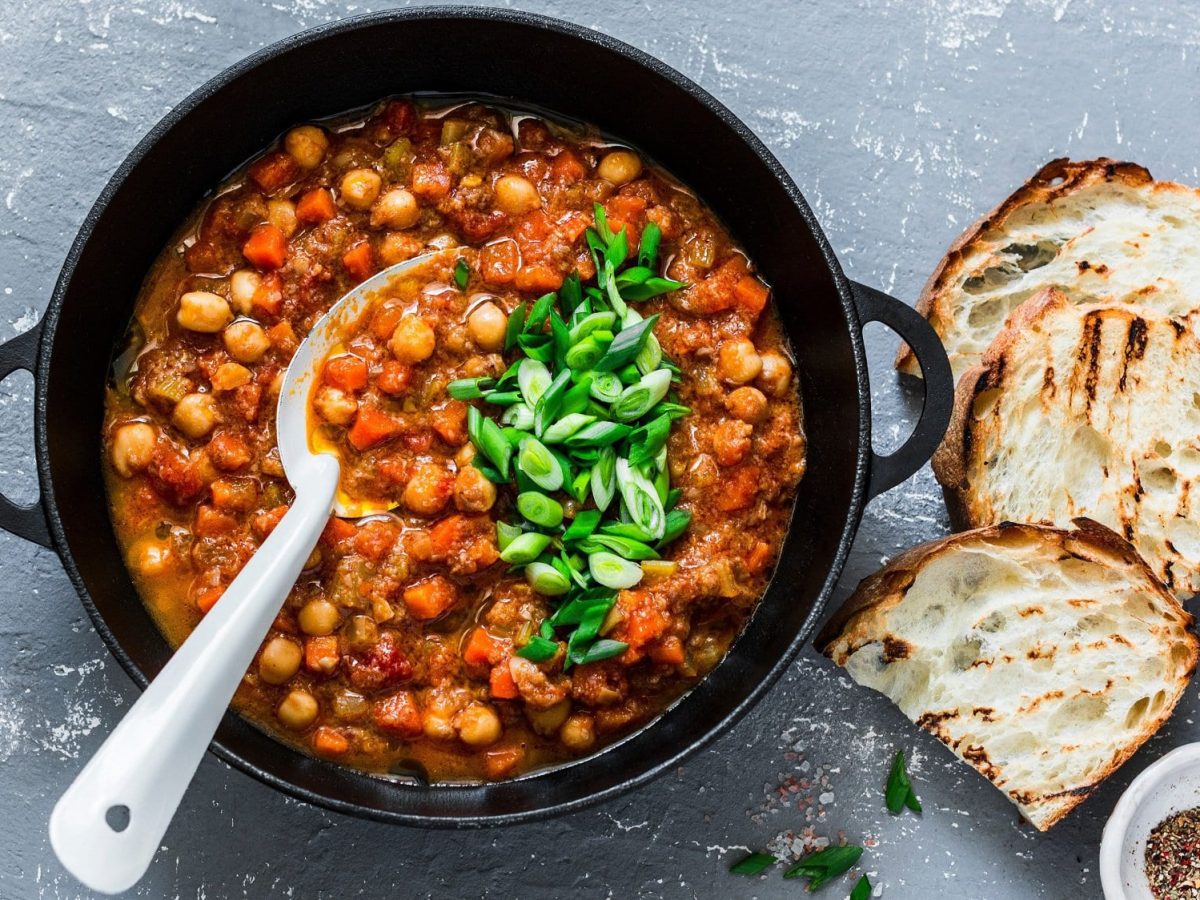 Ein Topf Kichererbsen-Chili mit Möhren, Tomaten und anderem Gemüse, außerdem Frühlingszwiebeln, daneben geröstete Brotscheiben und Schalen mit Gewürzen. Draufsicht.