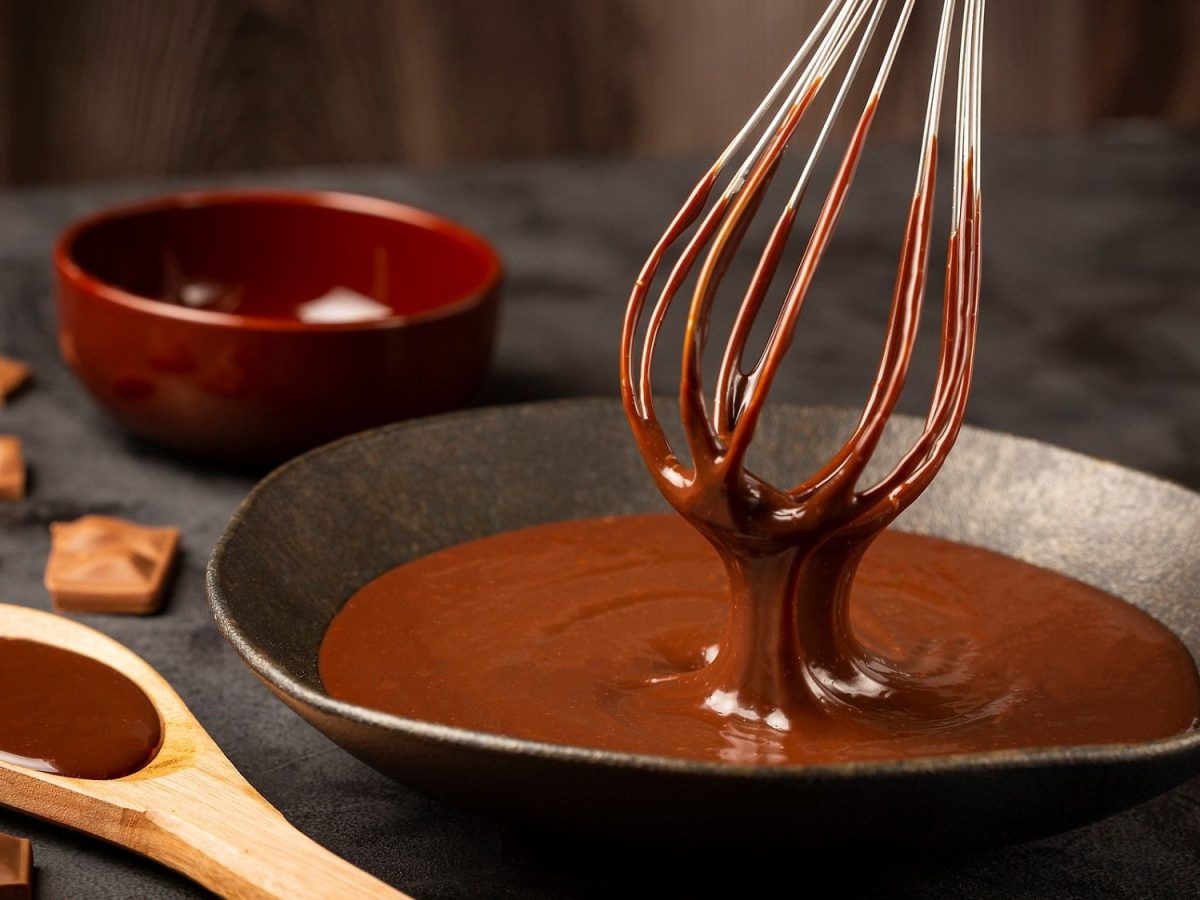 eine dunkler hintergrund, fronatlsicht. eine pfanne mit einer nougatsauve und einem schneebesen. daneben liegen schokolade und ein holzlöffel auf dem eine schokoladige sauce ist.