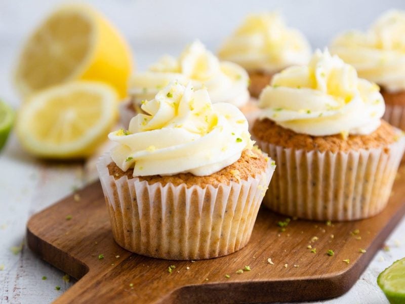 Ein paar Kokos-Limetten-Cupcakes auf einem Holzbrett, drumherum halbierte Limetten und Zitronen.