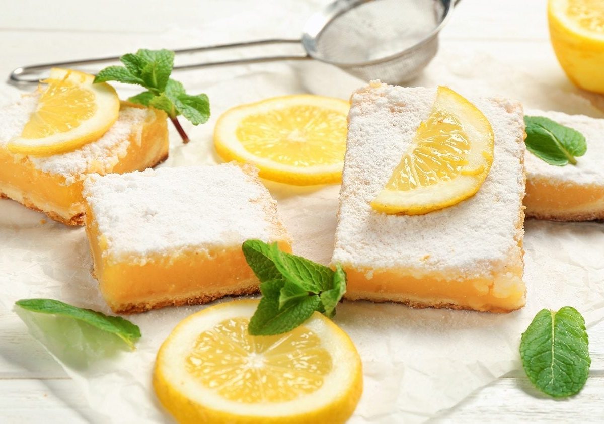 Frontalsicht: auf einem weißen Untergrund aus Holz liegen mehrere Lemon Bars bzw. Zitronenschnitten, Zitronenscheiben und Minzbätter.