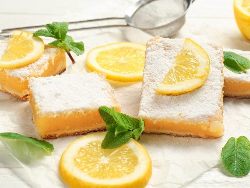 Frontalsicht: auf einem weißen Untergrund aus Holz liegen mehrere Lemon Bars bzw. Zitronenschnitten, Zitronenscheiben und Minzbätter.