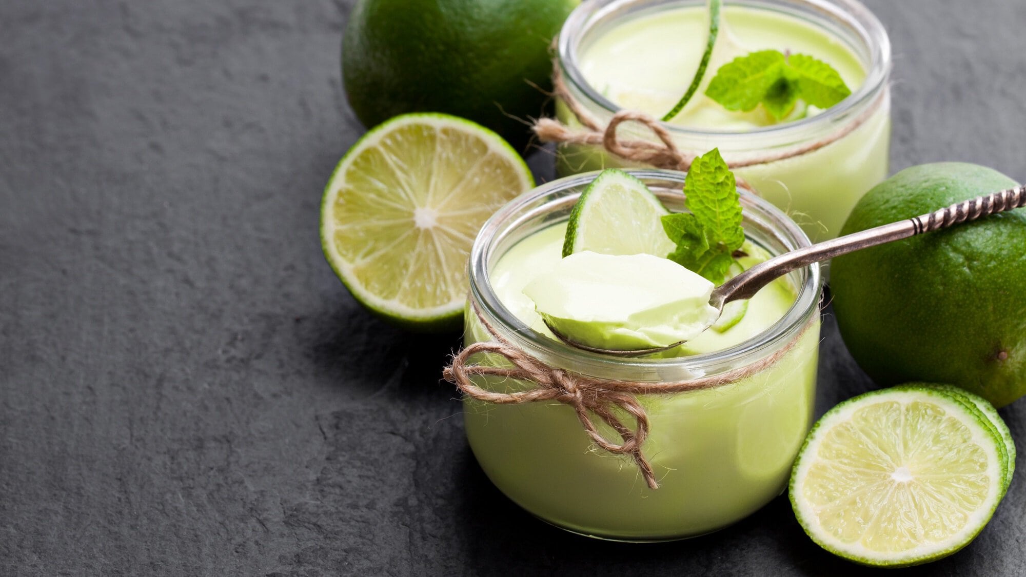 Draufsicht: Auf einer dunklen Holzplatte stehen zwei Gläser Limetten-Minz-Joghurt. Drumherum liegen ganze Limetten sowie Limettenscheiben.